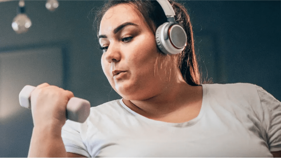 Medium shot of a woman wearing headphones while exercising, performing a bicep curl with a dumbbell.